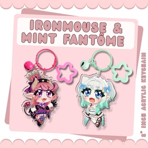 May include: Two 7.62 cm acrylic keychains featuring Ironmouse and Mint Fantôme characters. The Ironmouse charm has a pink ring, bell, and star clasp. The Mint Fantôme charm has a mint green ring, bell, and star clasp. Text reads: IRONMOUSE & MINT FANTÔME.