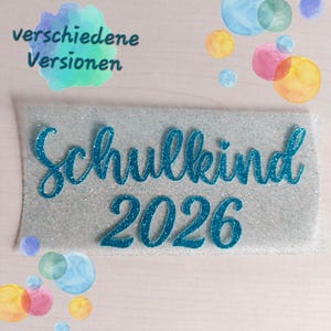 Könnte beinhalten: Eine rechteckige silberne Glitzerplatte mit dem blauen Glitzerschriftzug Schulkind 2026 in Schreibschrift. Der Hintergrund ist mit verschiedenen bunten Aquarellkreisen und dem Text verschiedene Versionen verziert.