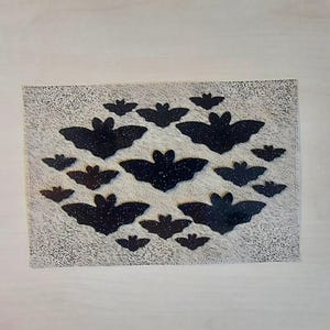 Könnte beinhalten: Ein rechteckiges Dekorationselement mit einem Muster aus schwarzen Fledermäusen mit Glitzerakzenten auf einem strukturierten, cremefarbenen Hintergrund. Die Fledermäuse variieren in der Größe und sind in einem sich wiederholenden Muster angeordnet.