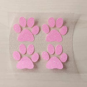 Könnte beinhalten: Vier rosa glitzernde Pfotenabdruck-Aufkleber auf einer durchsichtigen Folie.