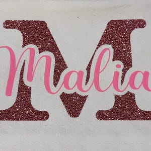 Könnte beinhalten: Eine weiße Canvas-Tragetasche mit einem großen pinkfarbenen Glitzerbuchstaben "M" und dem Namen "Malia" in pinkfarbenem Glitzer.