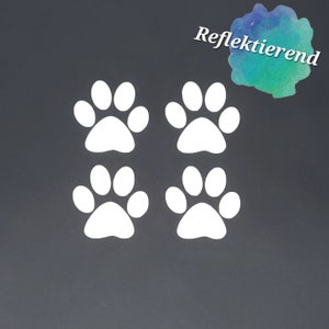 Könnte beinhalten: Vier weiße reflektierende Pfotenabdruck-Aufkleber auf einem dunkelgrauen Hintergrund. Das Wort "Reflektierend" ist in einer hellblauen, grünen und violetten Aquarell-Schriftart geschrieben.