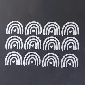 Könnte beinhalten: Eine Sammlung weißer Aufkleber in Regenbogenform mit Herzdetails auf schwarzem Hintergrund. Die Aufkleber sind in Reihen und Spalten angeordnet und erzeugen ein sich wiederholendes Muster. Das Design ist schlicht und minimalistisch.