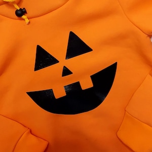 Könnte beinhalten: Orangefarbener Hoodie mit einem schwarzen Kürbisgesicht, das auf der Vorderseite aufgedruckt ist.