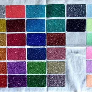 Könnte beinhalten: Eine Sammlung rechteckiger Glitzerproben in verschiedenen Farben, darunter Silber, Rot, Blau, Grün, Gold und Rosa. Die Proben sind auf einem weißen Stoffhintergrund angeordnet und zeigen die Auswahl an Glitzeroptionen.