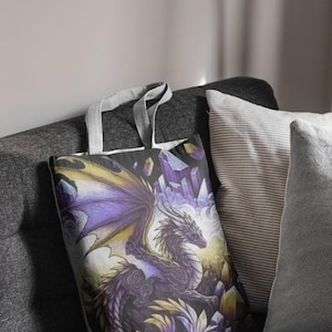 May include: A fabric tote bag featuring a detailed illustration of a purple and yellow dragon surrounded by jagged crystals. The dragon has intricate scale patterns and large wings, set against a backdrop of glowing light and crystalline formations.