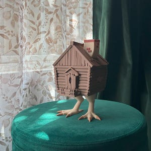 May include: A whimsical brown cabin sculpture with a chimney, perched on realistic chicken legs. The cabin features log-cabin style construction and a small door. The sculpture is displayed on a green velvet surface.