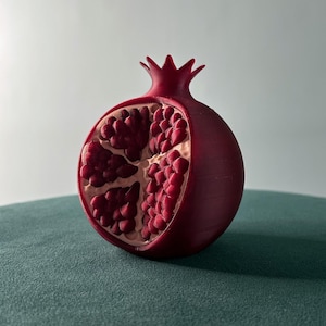 Peut inclure: Une grenade rouge en 3D imprimée, coupée en deux, révélant les graines à l'intérieur. La grenade est posée sur une surface verte.