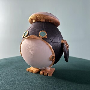 May include: A 3D printed, white and black penguin figurine with gold accents. The penguin has a round body and a small beak. It is wearing a gold and white hat.