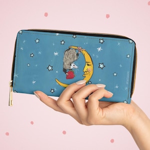 May include: A blue zip-around wallet featuring a whimsical design of a possum hanging from a crescent moon with a smiling face, surrounded by stars. The wallet has a gold zipper and a black trim.