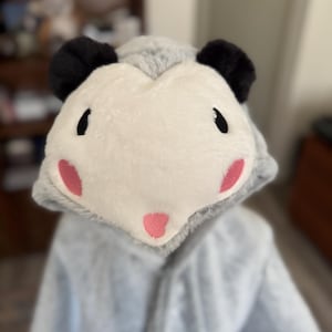 May include: A plush gray hooded garment featuring an opossum face design on the hood. The hood has a white face patch, black ears, embroidered black eyes, and pink accents on the cheeks and nose, crafted from soft, fuzzy material.