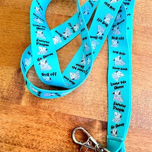 May include: A turquoise lanyard with a repeating pattern of a cartoon animal and phrases like "Bug Off" and "Leave Me Alone." The lanyard has a silver-colored metal clasp at the end. The text and animal are white and gray.