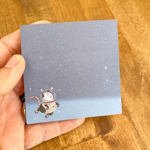 May include: A square notepad featuring a cartoon opossum in a space suit against a starry, blue-gray background. The opossum is depicted with a pink tail and is wearing a white helmet with a red collar. The notepad is held in a hand.