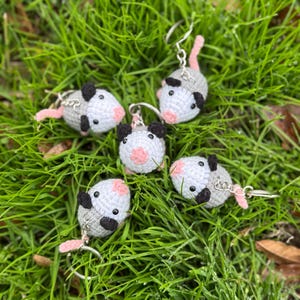 May include: Five handmade crochet possum keychains. Each possum has a gray body, white face, black ears, and a pink nose and tail. The keychains are lying on green grass.