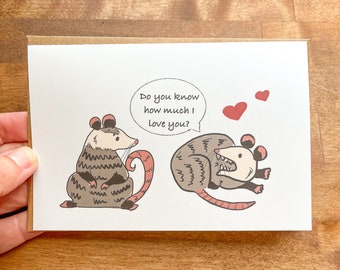 May include: A greeting card with a cartoon illustration of two opossums. One opossum is holding a speech bubble that says "Do you know how much I love you?" The other opossum is holding a heart in its mouth. The card is on a wooden surface.