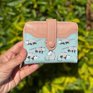 May include: A small, bi-fold wallet with a light blue background featuring a repeating pattern of opossums. The wallet has a tan leather flap and a snap closure. The zipper is visible on the side.