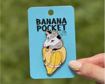 May include: A metal enamel pin featuring a gray and white opossum peeking out of a yellow banana. The pin is attached to a light blue rectangular display card with the text BANANA POCKET POSSUM printed in black lettering at the top.
