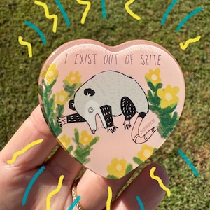 May include: A heart-shaped, pink trinket featuring a cartoon opossum illustration. The opossum is lying down with its arms outstretched, surrounded by yellow flowers. Text reads "I EXIST OUT OF SPITE". Decorative yellow and blue lines surround the trinket.