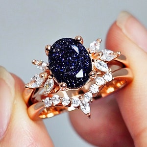 Peut inclure: Une bague en or rose avec une pierre centrale ovale en grès bleu foncé aux inclusions scintillantes. La monture est ornée de plusieurs pierres claires taille marquise disposées en grappe florale, accompagnée d'une alliance en V assortie.