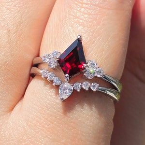 May include: A silver ring with a red gemstone and smaller clear gemstones. The ring is on a finger.