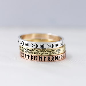 May include: Three stacked silver, gold, and rose gold rings with different engraved designs. The top ring has a repeating pattern of moons, stars, and suns. The middle ring has a repeating pattern of eyes. The bottom ring has a repeating pattern of runes.