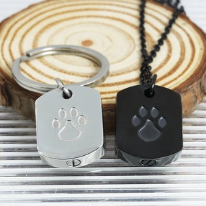 May include: Two dog paw print cremation urns, one silver and one black. The urns are shaped like dog tags and have a screw-on lid. The silver urn is attached to a key ring and the black urn is attached to a black chain.