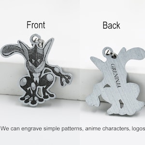 May include: A metal pendant featuring a detailed engraving of a Greninja character from the front and back. The front shows the character's face and body, while the back has the word "GRENINJA". The pendant has a small loop for attaching to a chain or keychain.