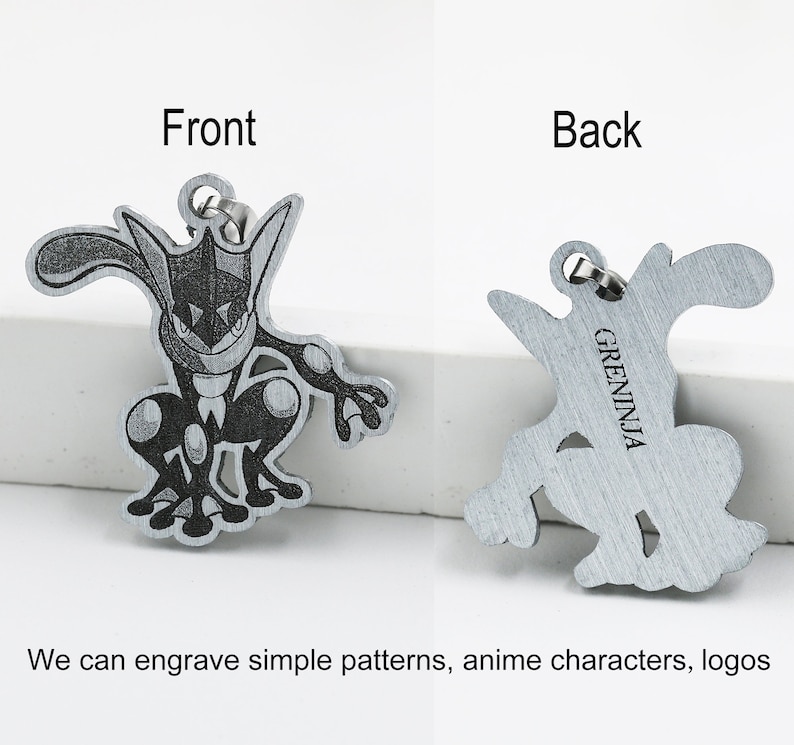 May include: A metal pendant featuring a detailed engraving of a Greninja character from the front and back. The front shows the character's face and body, while the back has the word "GRENINJA". The pendant has a small loop for attaching to a chain or keychain.