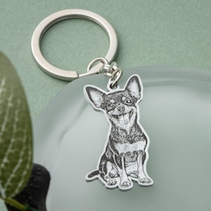 May include: A silver-colored keychain featuring a smiling Chihuahua dog illustration. The dog is black and white, with its tongue sticking out. The keychain has a round ring for attaching keys.