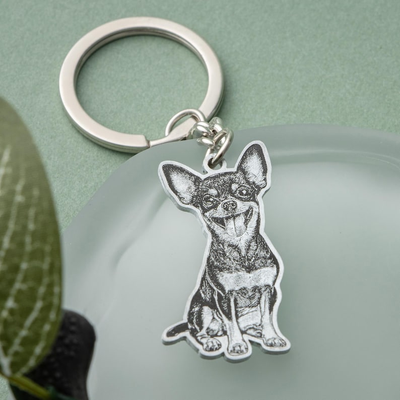 May include: A silver-colored keychain featuring a smiling Chihuahua dog illustration. The dog is black and white, with its tongue sticking out. The keychain has a round ring for attaching keys.