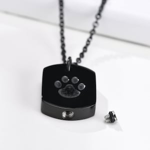 May include: A black chain necklace with a black rectangular pendant. The pendant has a silver paw print engraved on it. The pendant has a small opening on the bottom, and a separate silver screw top is shown next to it.