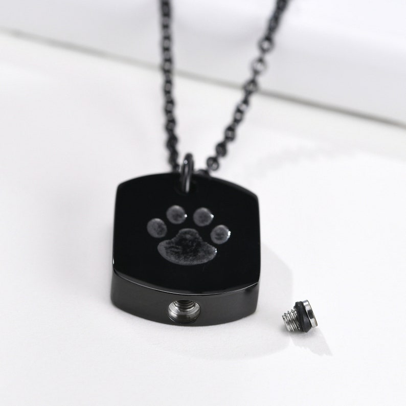 May include: A black chain necklace with a black rectangular pendant. The pendant has a silver paw print engraved on it. The pendant has a small opening on the bottom, and a separate silver screw top is shown next to it.
