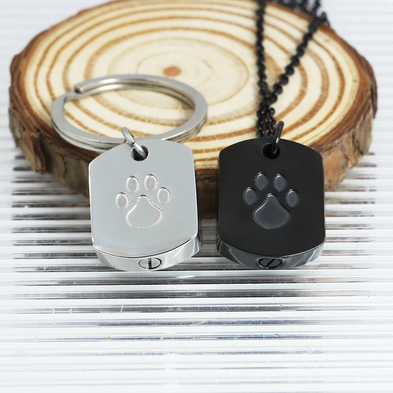 May include: Two dog paw print cremation urns, one silver and one black. The urns are shaped like dog tags and have a screw-on lid. The silver urn is attached to a key ring and the black urn is attached to a black chain.