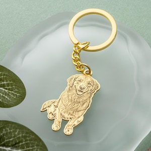 May include: A gold-colored keychain featuring a detailed engraving of a dog. The dog is depicted in a sitting position with a smiling expression. The keychain includes a chain and a circular ring for attaching keys. The background is a light blue surface.
