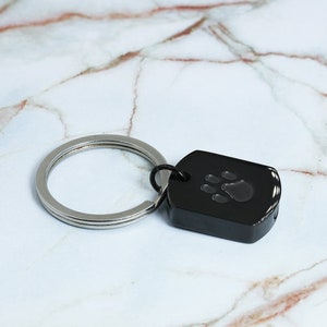 May include: A black metal keychain with a silver ring and a paw print design. The keychain is on a white marble surface.