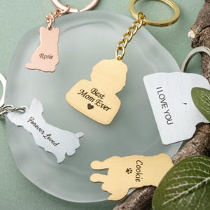 May include: A collection of personalized keychains in various shapes and colors. Keychains include a gold-colored silhouette with "Best Mom Ever" text, a silver dog-shaped keychain with "Forever Loved", and a gold-colored dog-shaped keychain with "Cookie".
