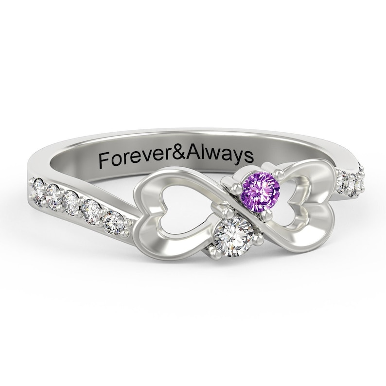May include: A silver ring with two hearts intertwined to form an infinity symbol. The ring is set with two gemstones, one purple and one white, and has a row of smaller white gemstones along the band. The inscription "Forever&Always" is engraved on the inside of the band.