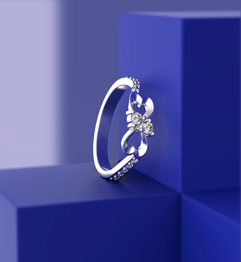 May include: A silver ring with a delicate design featuring two intertwined hearts and three small diamonds. The ring is displayed on a blue background.
