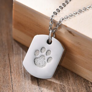 May include: A silver dog tag necklace with a paw print design. The tag is rectangular and has a smooth, polished finish. The chain is silver and has a delicate, link-style design.