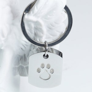 May include: A silver metal keychain with a silver metal tag. The tag is shaped like a rectangle with rounded corners and has a paw print design engraved in the center.