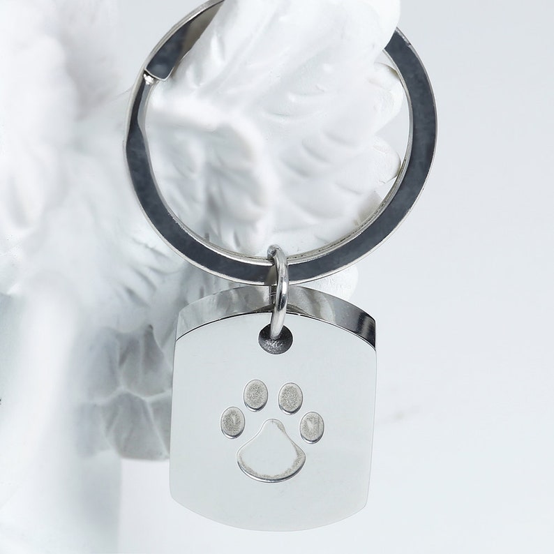 May include: A silver metal keychain with a silver metal tag. The tag is shaped like a rectangle with rounded corners and has a paw print design engraved in the center.