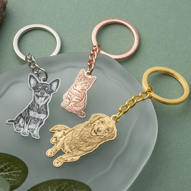 May include: Three metal keychains featuring engraved pet portraits: a Chihuahua, a tabby cat, and a golden retriever. Each keychain has a different metal finish: silver, rose gold, and gold. The keychains are connected to a ring with a chain.