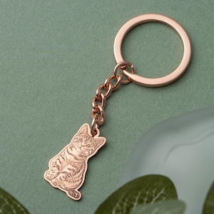 May include: A rose gold cat-shaped keychain. The keychain features a detailed engraving of a seated cat, connected to a chain and a circular key ring. The background is a muted green, with a hint of a blurred object.