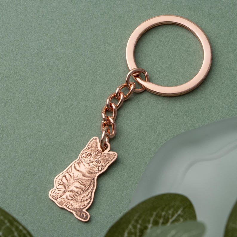 May include: A rose gold cat-shaped keychain. The keychain features a detailed engraving of a seated cat, connected to a chain and a circular key ring. The background is a muted green, with a hint of a blurred object.
