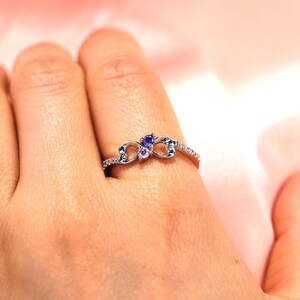 May include: A silver ring with two hearts intertwined, featuring a small purple gemstone and a smaller clear gemstone.