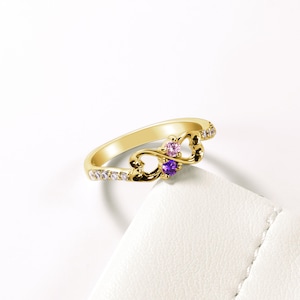 May include: Gold infinity ring with two small gemstones, one purple and one pink, set in a heart design. The ring is adorned with small clear stones.