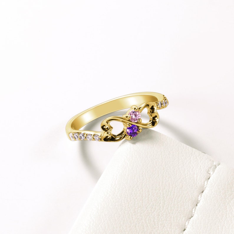 May include: Gold infinity ring with two small gemstones, one purple and one pink, set in a heart design. The ring is adorned with small clear stones.