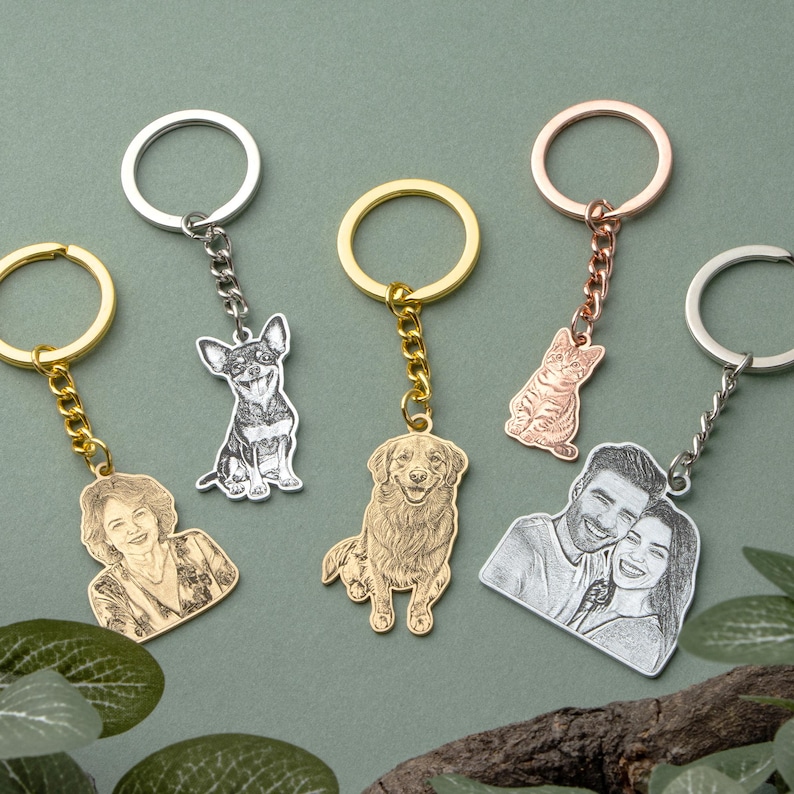 May include: A collection of personalized keychains featuring engraved portraits and pet designs. The keychains have various metal finishes, including silver, gold, and rose gold. The designs include a dog, a cat, a couple, and a portrait of a person.