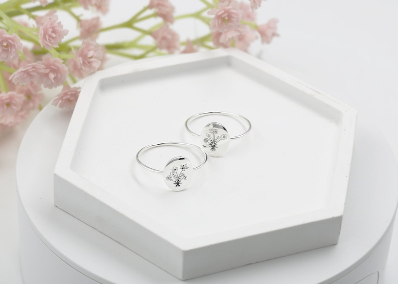 May include: Two silver rings with a floral design on the front. The rings are on a white hexagonal display stand.