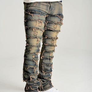 May include: Distressed blue jeans with multiple horizontal frayed sections. The jeans have a light wash and a relaxed fit. The model is wearing white sneakers. The jeans are a unique fashion statement.